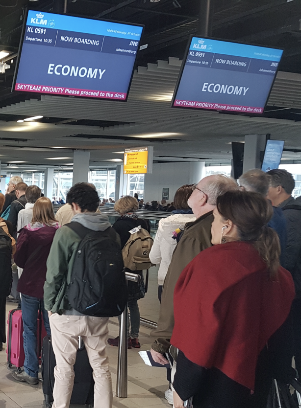 A man in a boarding queue looking up at a boarding screen. The screen says "Economy".